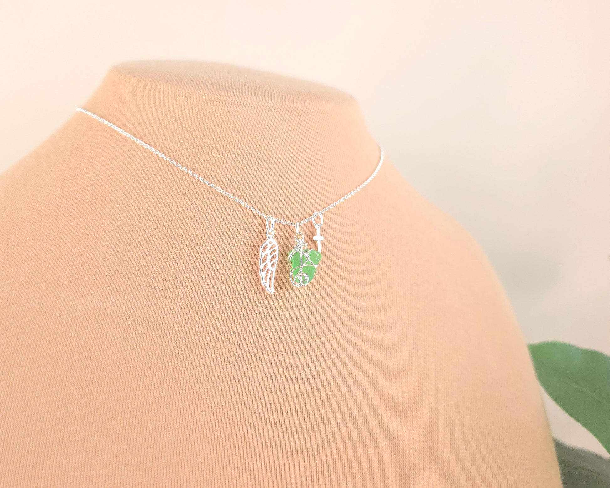Silver necklace with green sea glass wire wrapped pendant with a silver angel wing and small cross on chain with beige background