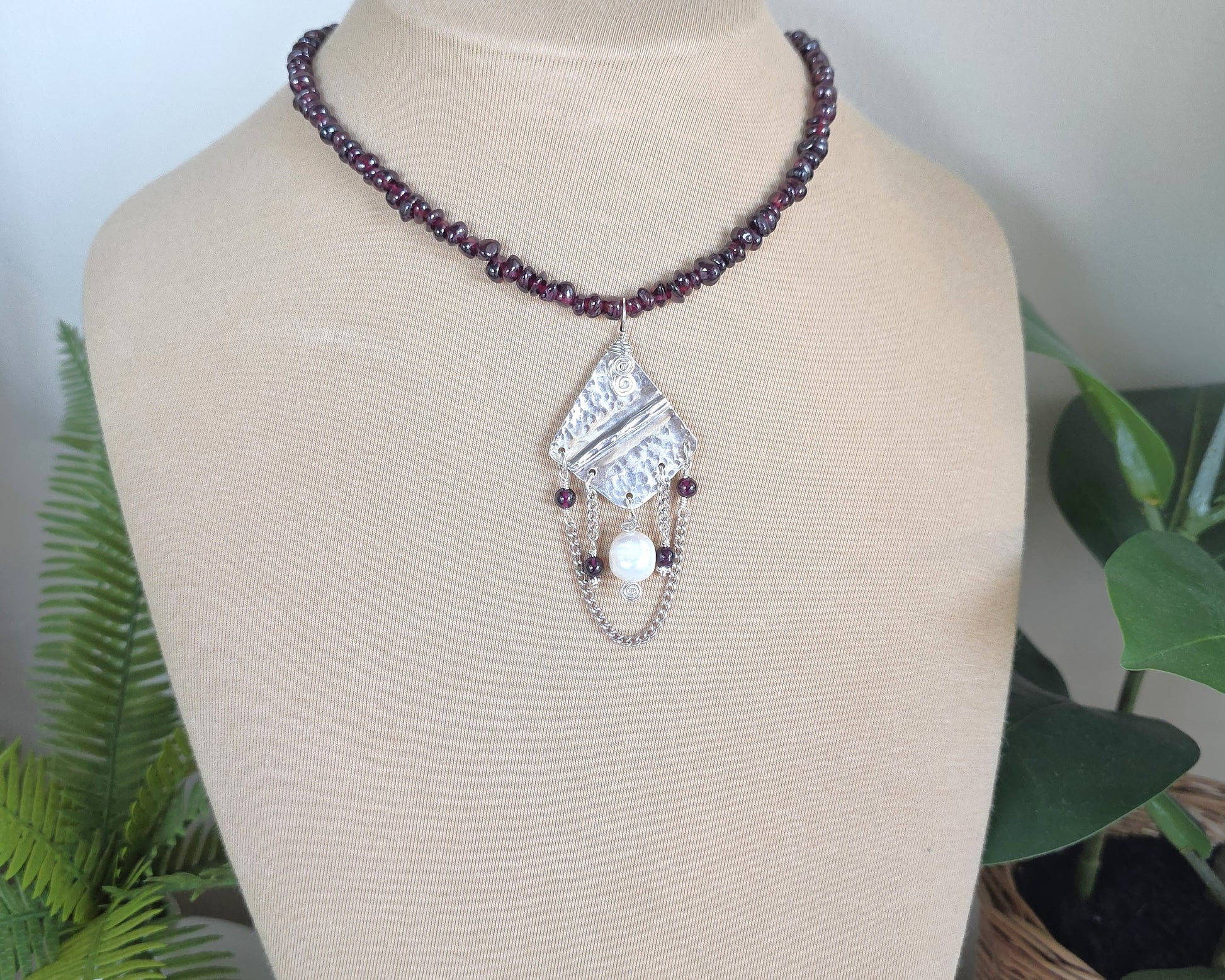 Deep Red stone beaded lariat style necklace with a Large Silver pendant with dangling stones white pearl and chains on beige  display bust and plants 