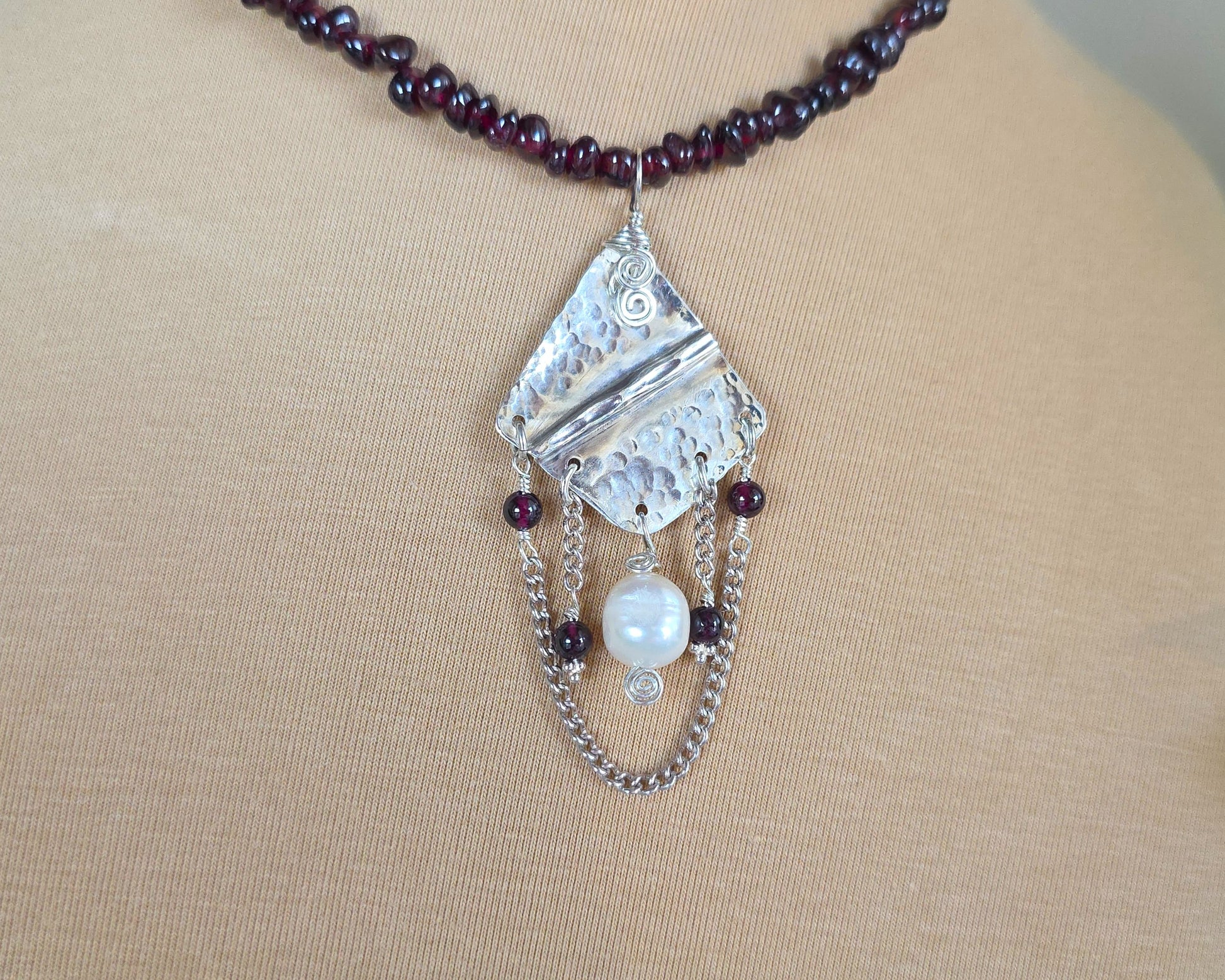 Deep Red stone beaded lariat style necklace with a Large Silver pendant with dangling stones white pearl and chains on beige  display bust and plants 