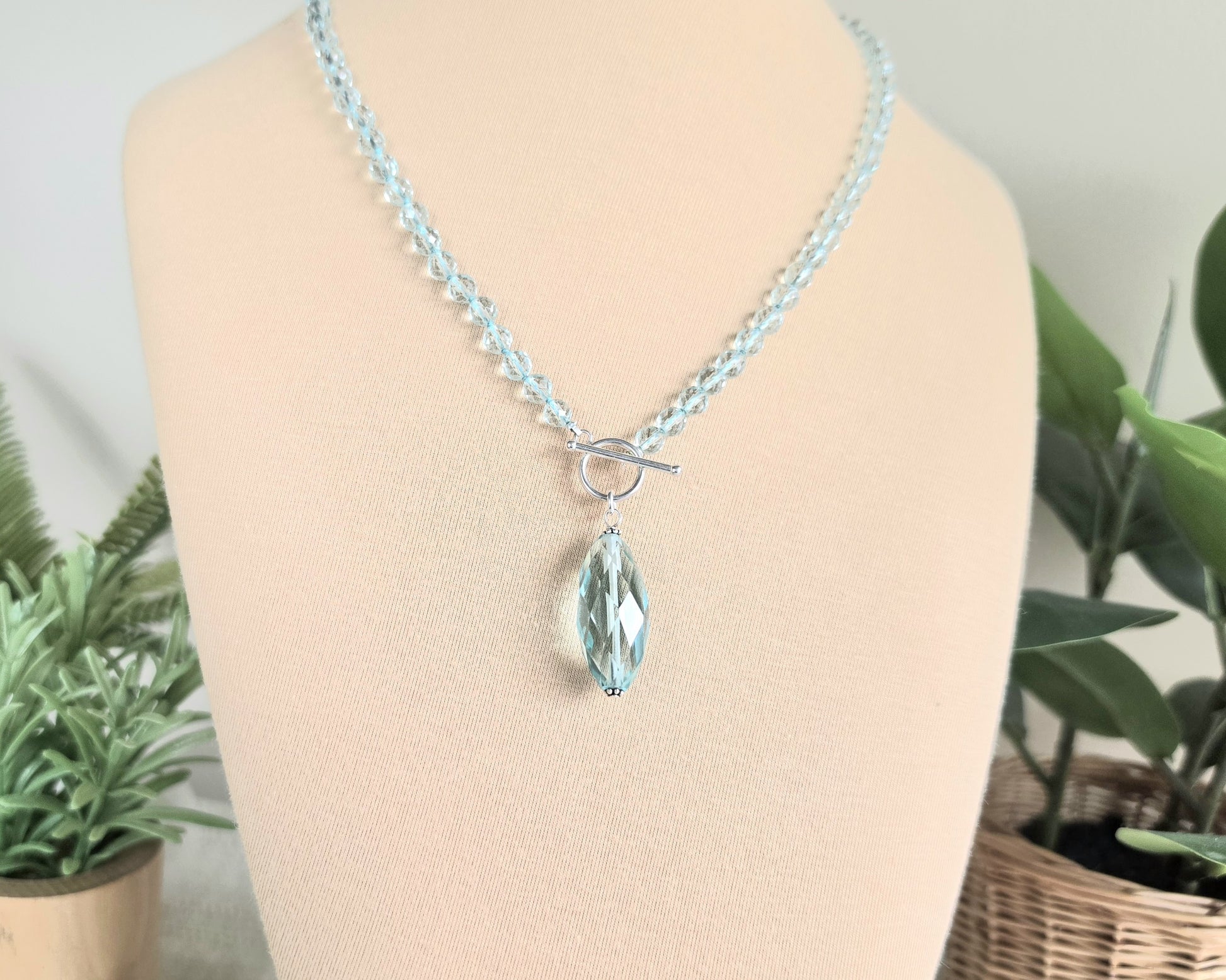 Sparkly Aquamarine hand knotted clear pale aqua blue faceted stone beaded necklace with silver toggle clasp and large stone pendant on beige display and plants in the background 