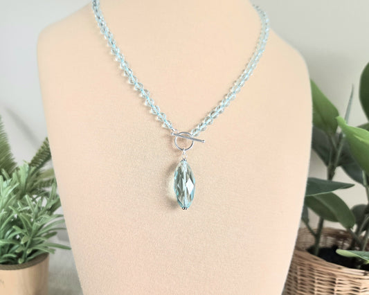 Sparkly Aquamarine hand knotted clear pale aqua blue faceted stone beaded necklace with silver toggle clasp and large stone pendant on beige display and plants in the background 