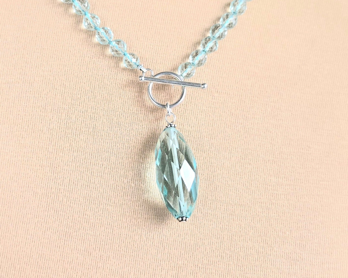 Sparkly Aquamarine hand knotted clear pale aqua blue faceted stone beaded necklace with silver toggle clasp and large stone pendant on beige  background 