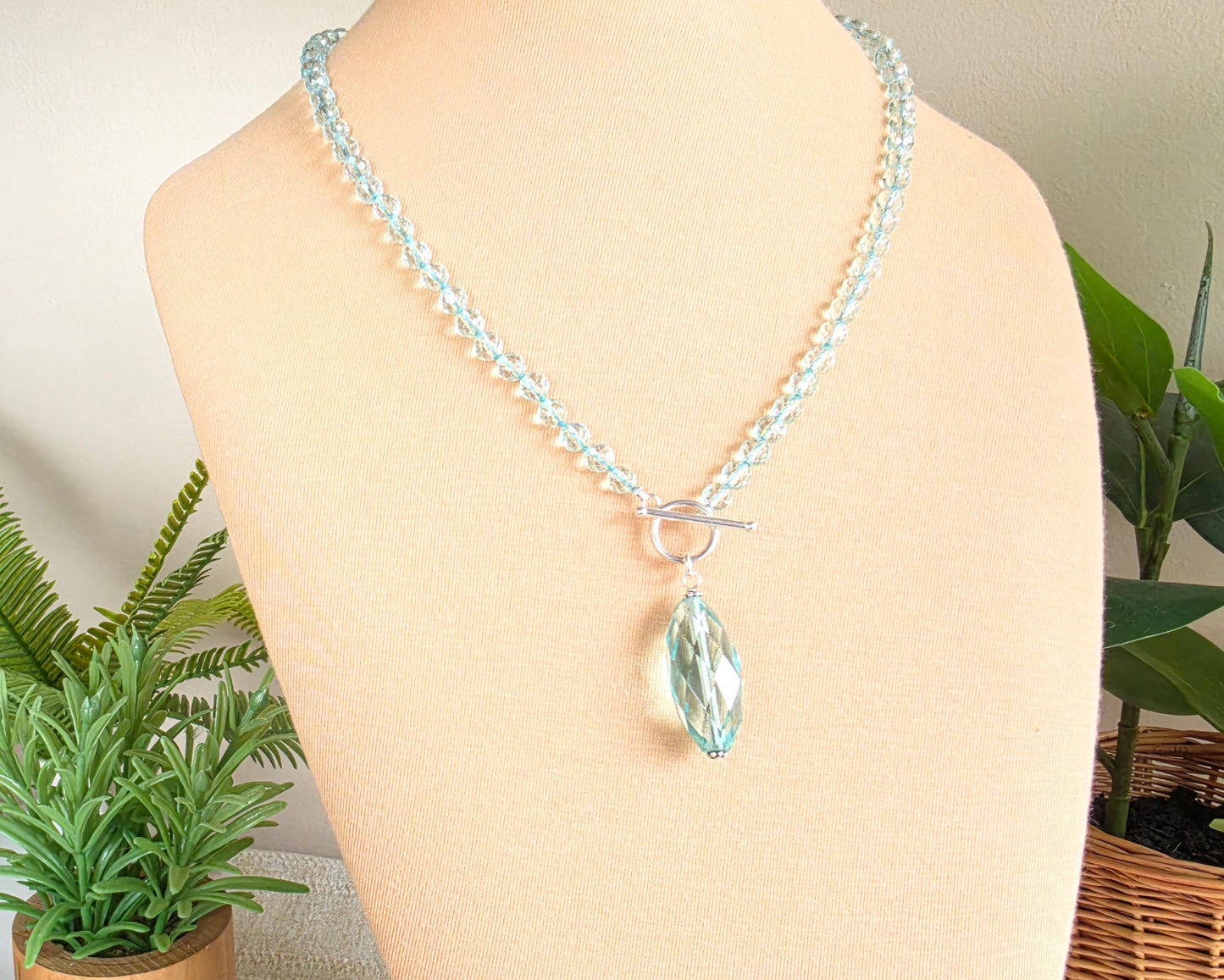 Sparkly Aquamarine hand knotted clear pale aqua blue faceted stone beaded necklace with silver toggle clasp and large stone pendant on beige display and plants in the background 