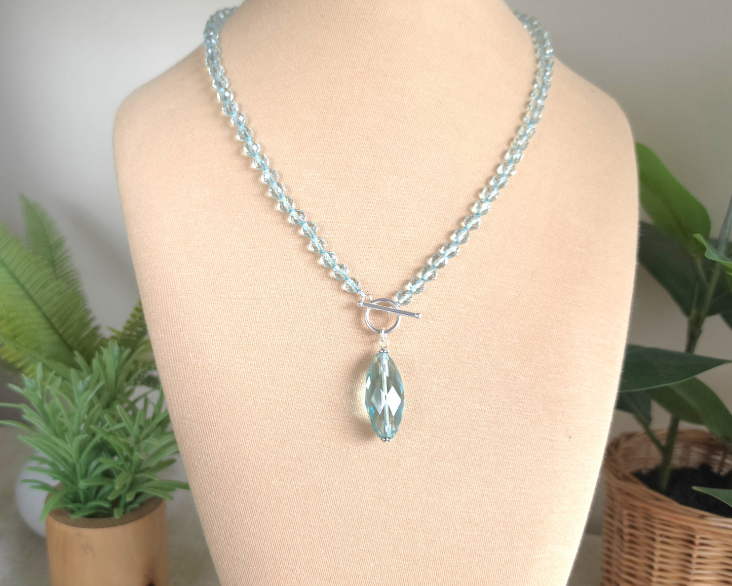 Sparkly Aquamarine hand knotted clear pale aqua blue faceted stone beaded necklace with silver toggle clasp and large stone pendant on beige display and plants in the background 