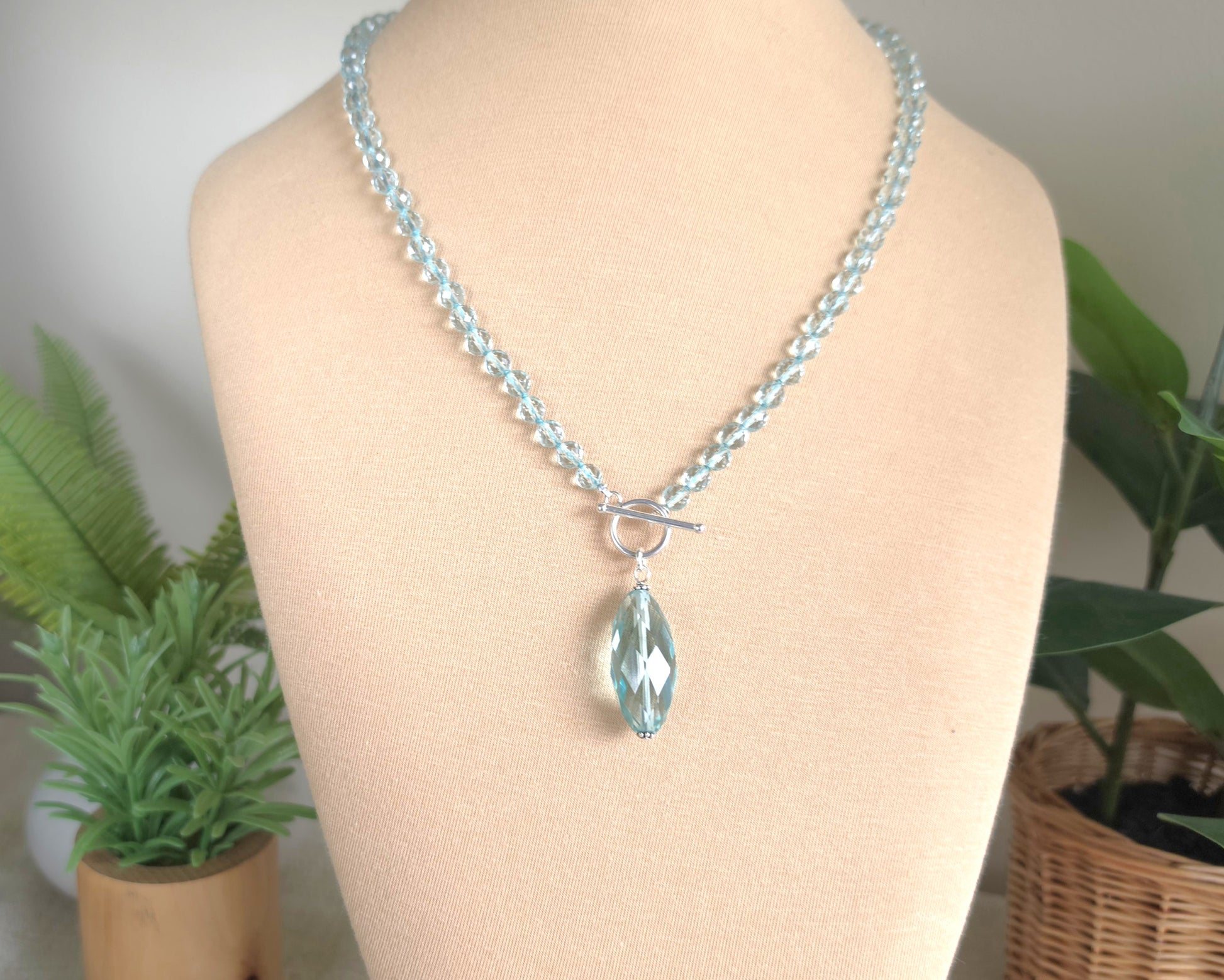 Sparkly Aquamarine hand knotted clear pale aqua blue faceted stone beaded necklace with silver toggle clasp and large stone pendant on beige display and plants in the background 