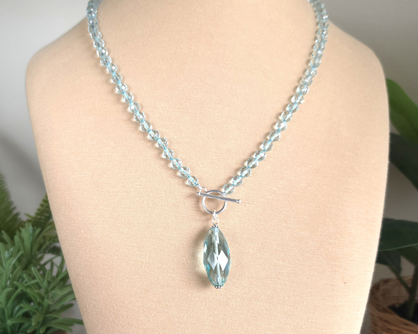 Sparkly Aquamarine hand knotted clear pale aqua blue faceted stone beaded necklace with silver toggle clasp and large stone pendant on beige display and plants in the background 
