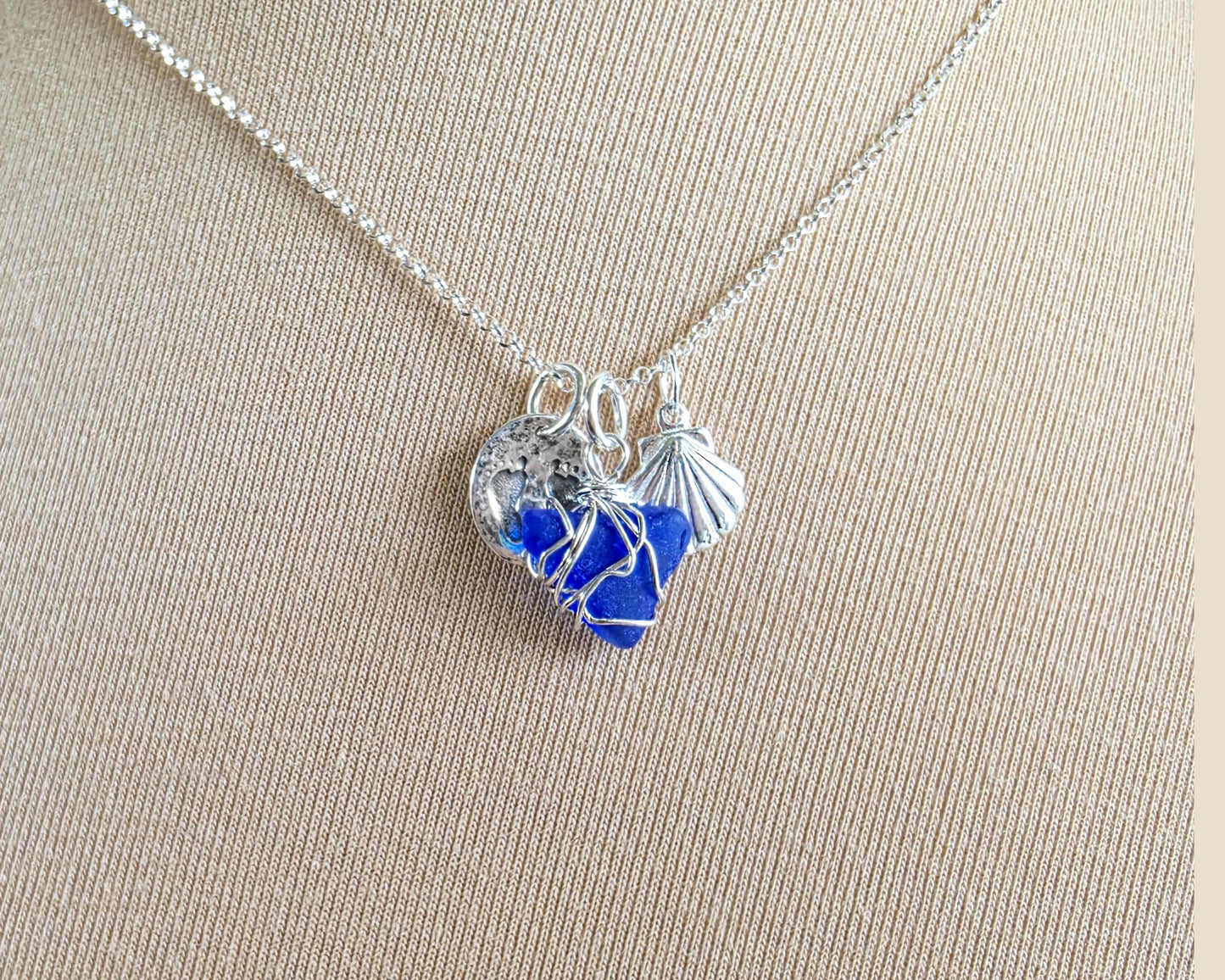 Sterling Silver three charm necklace with a deep blue sea glass wire wrapped pendant with shell and footprints pendants on chain on beige background 