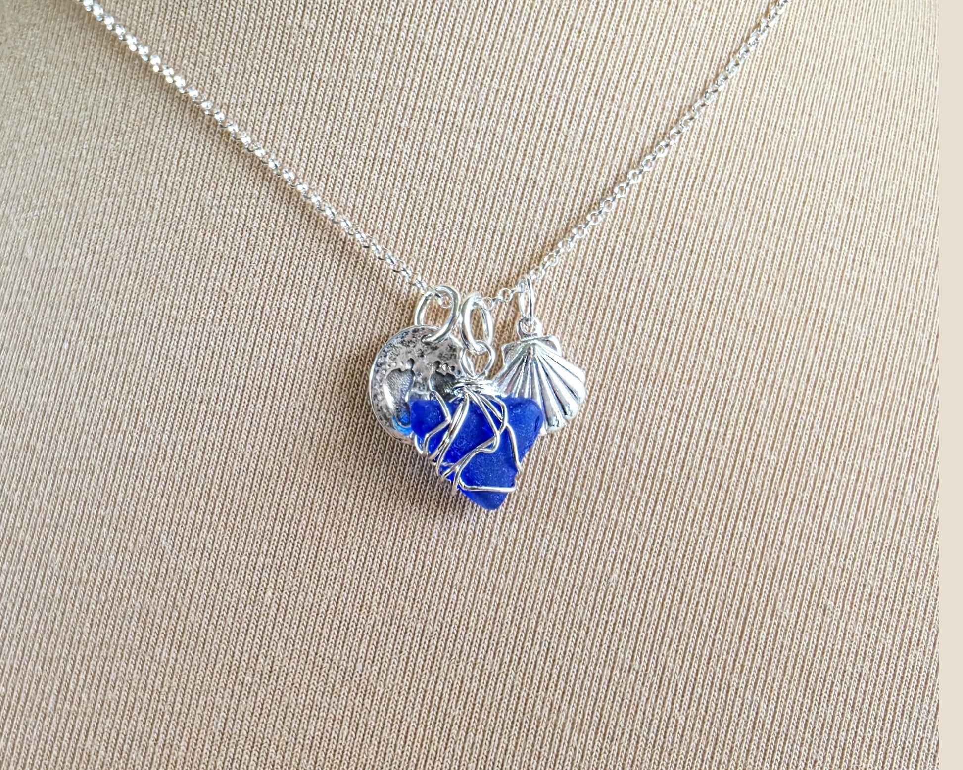 Sterling Silver three charm necklace with a deep blue sea glass wire wrapped pendant with shell and footprints pendants on chain on beige background 