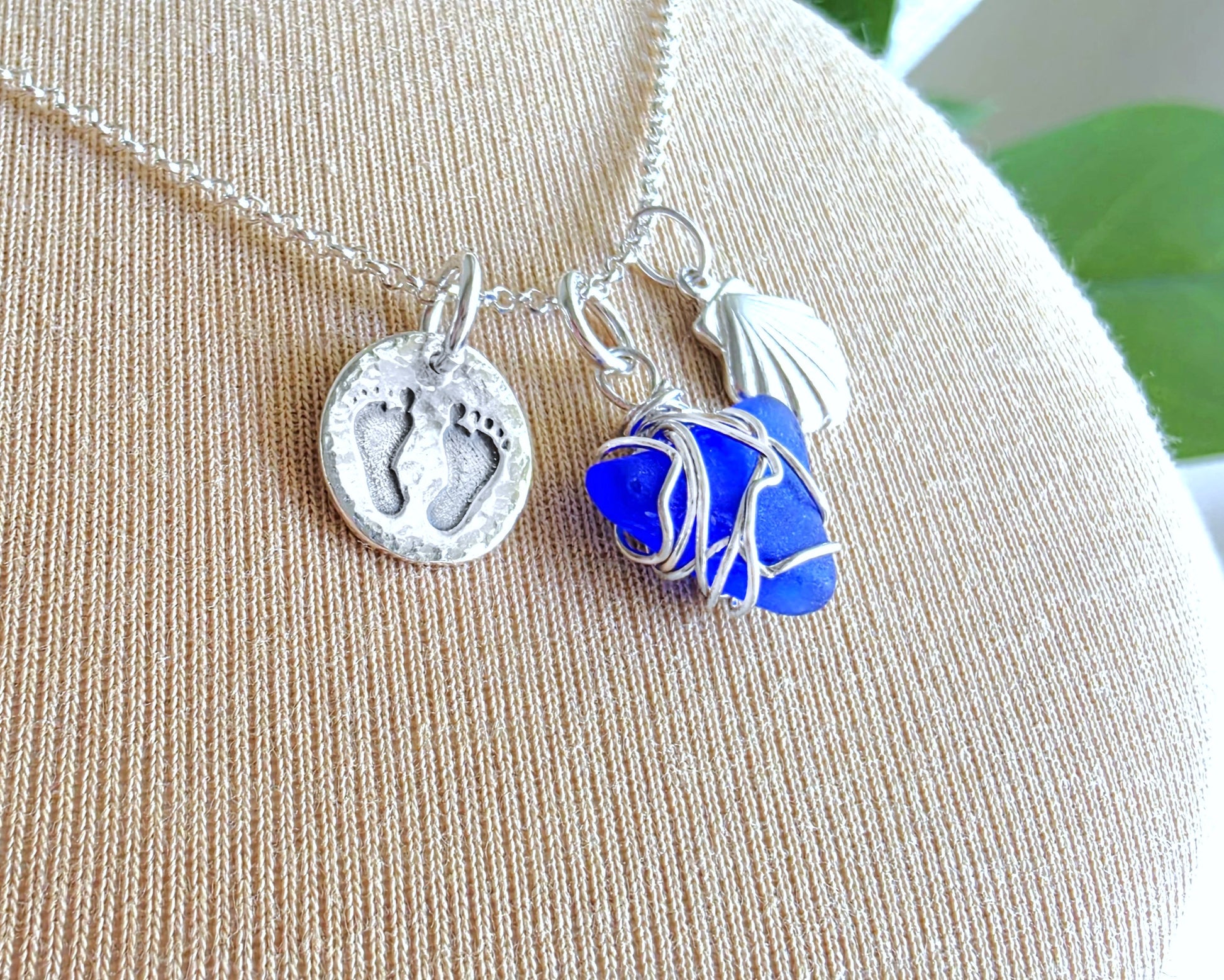 Sterling Silver three charm necklace with a deep blue sea glass wire wrapped pendant with shell and footprints pendants on chain on beige background 