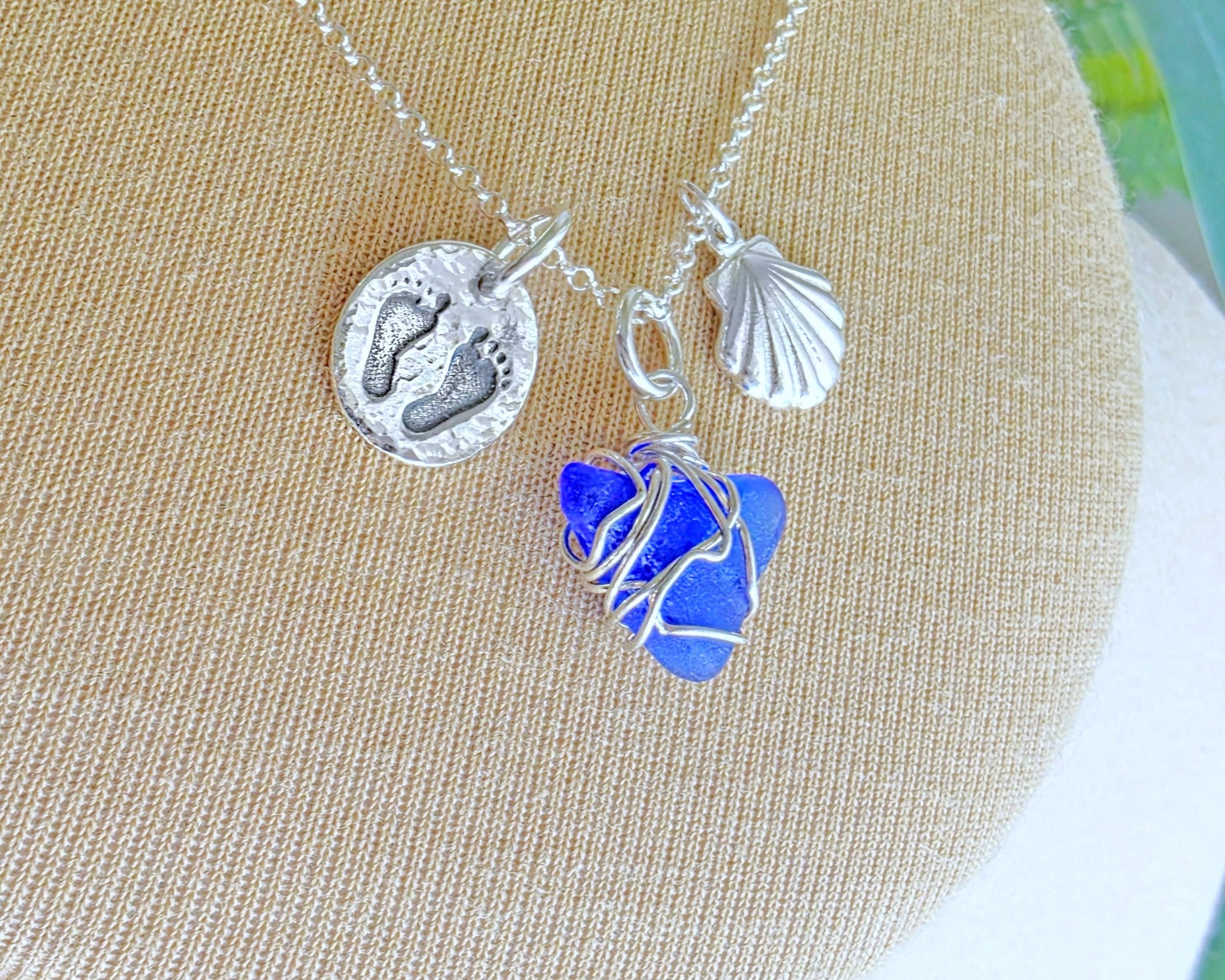 Sterling Silver three charm necklace with a deep blue sea glass wire wrapped pendant with shell and footprints pendants on chain on beige background 