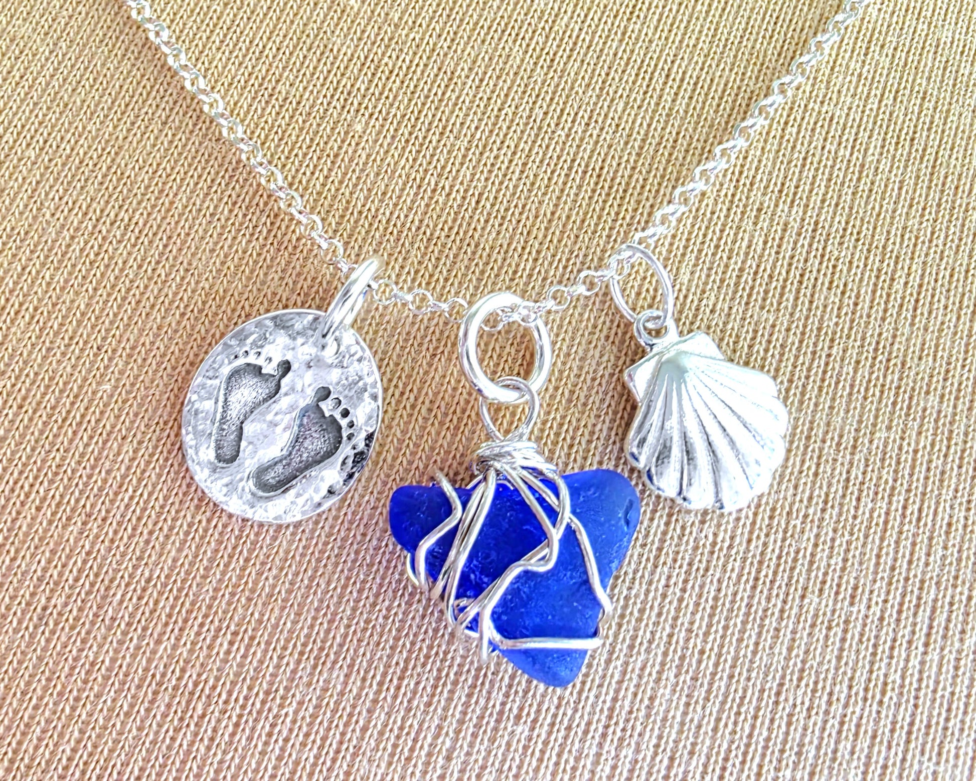 Sterling Silver three charm necklace with a deep blue sea glass wire wrapped pendant with shell and footprints pendants on chain on beige background 