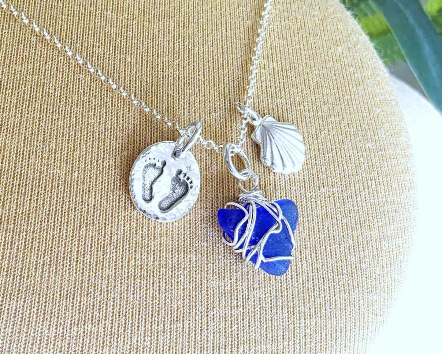 Sterling Silver three charm necklace with a deep blue sea glass wire wrapped pendant with shell and footprints pendants on chain on beige background 