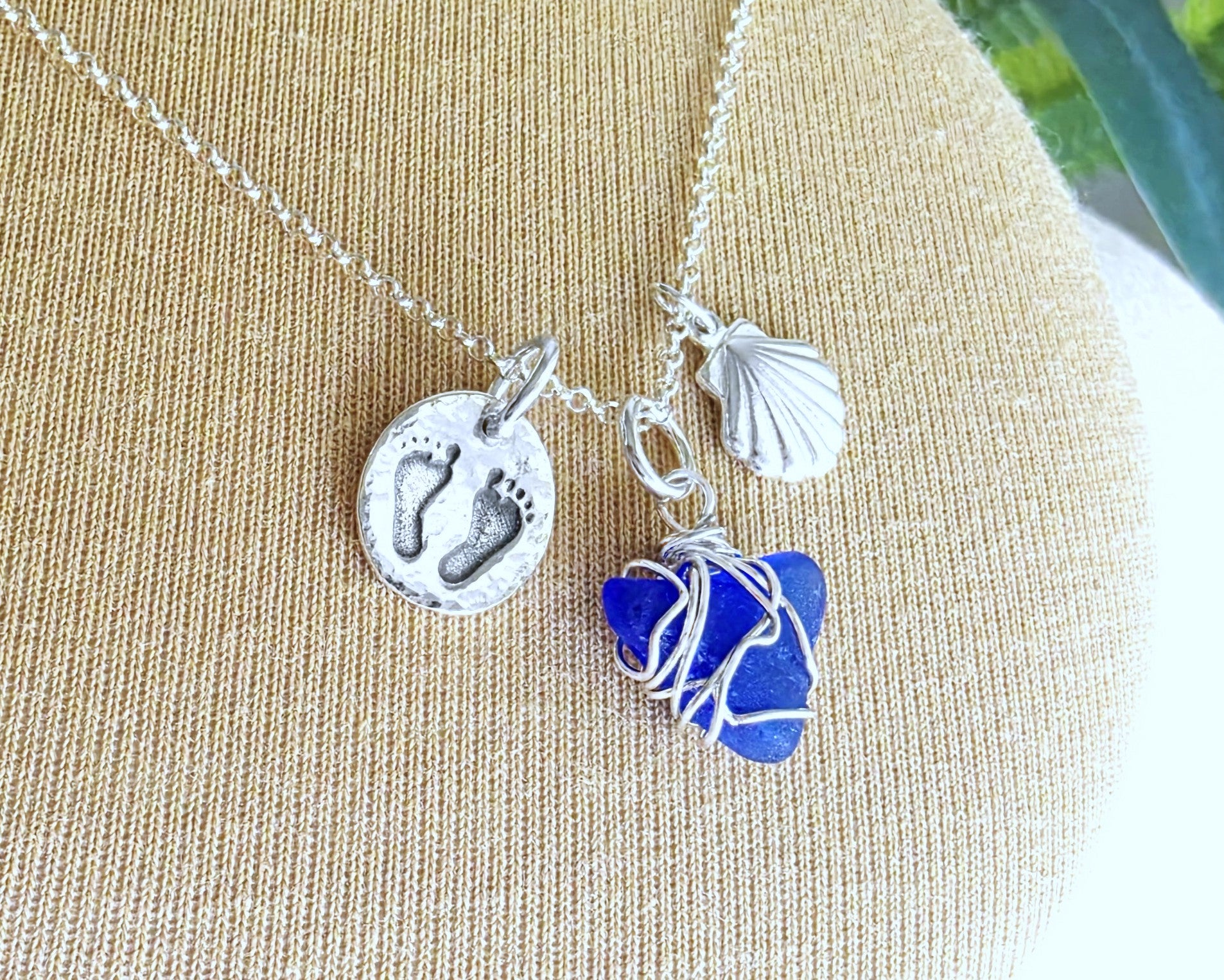 Sterling Silver three charm necklace with a deep blue sea glass wire wrapped pendant with shell and footprints pendants on chain on beige background 