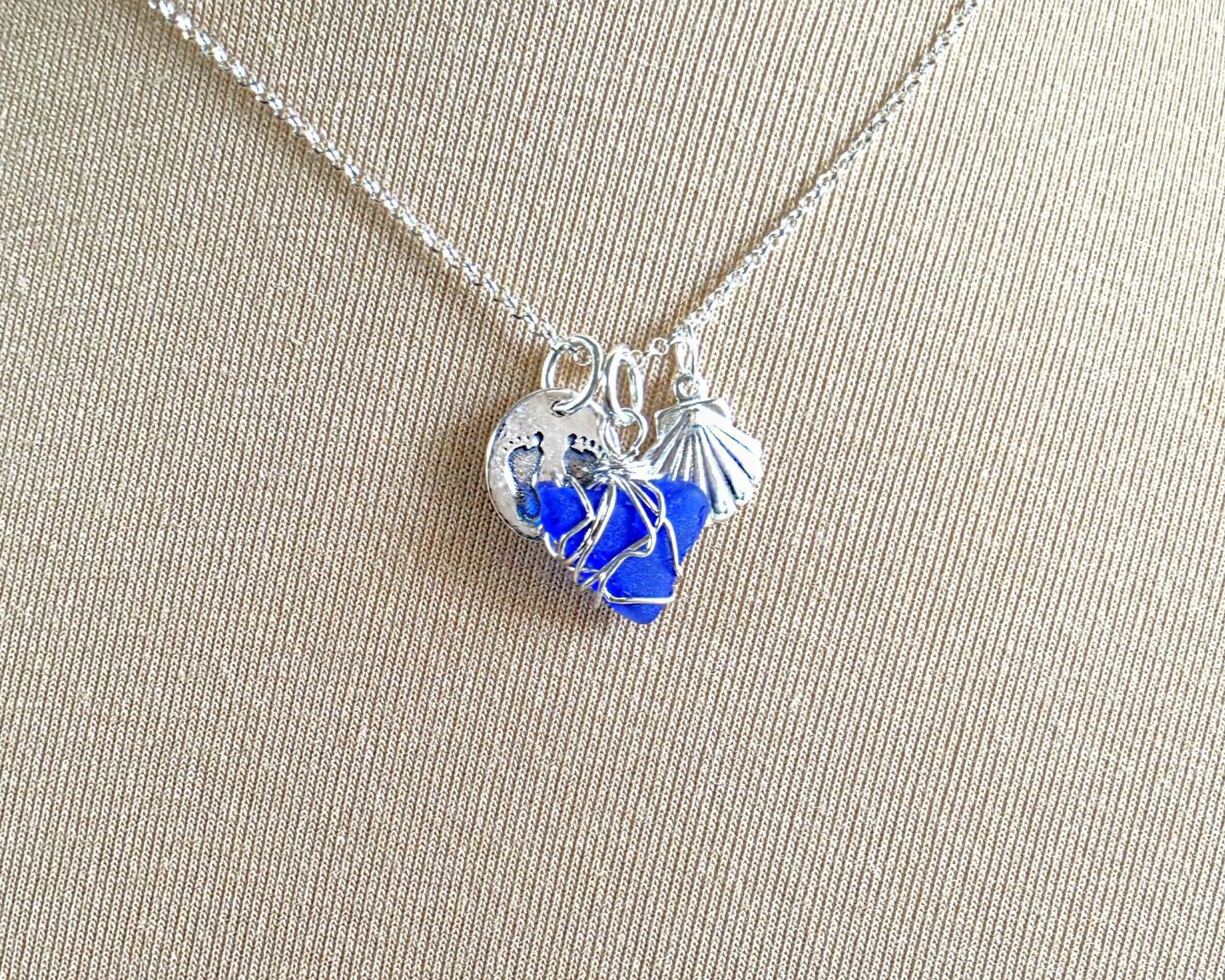 Sterling Silver three charm necklace with a deep blue sea glass wire wrapped pendant with shell and footprints pendants on chain on beige background 