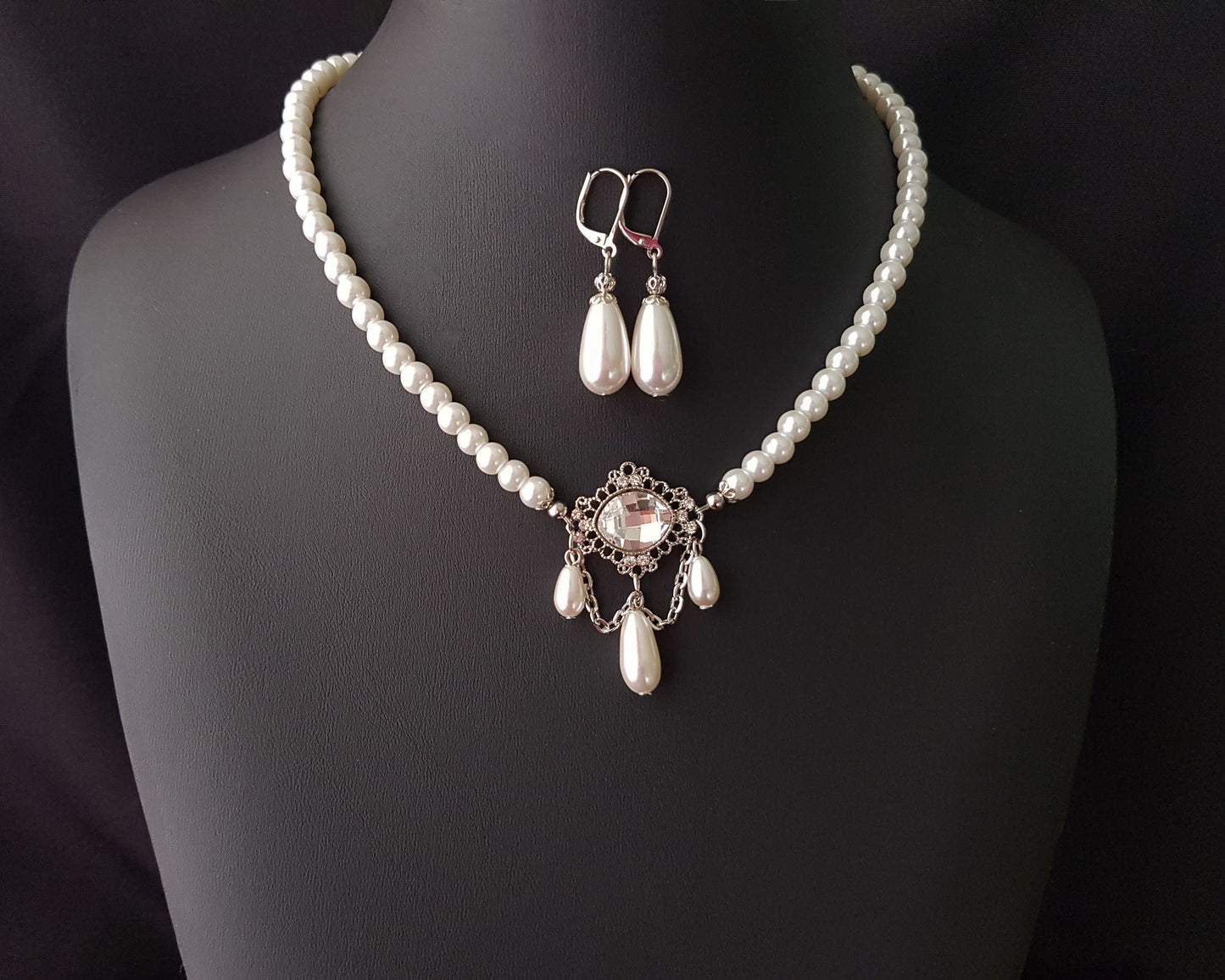Victorian Style Pearl Crystal Necklace and Earrings Set with white pearl necklace with large clear crystal and three dangling drop pearls and long back chain with another pearl drop. The matching earrings are drop shaped pearls, displayed on black. 