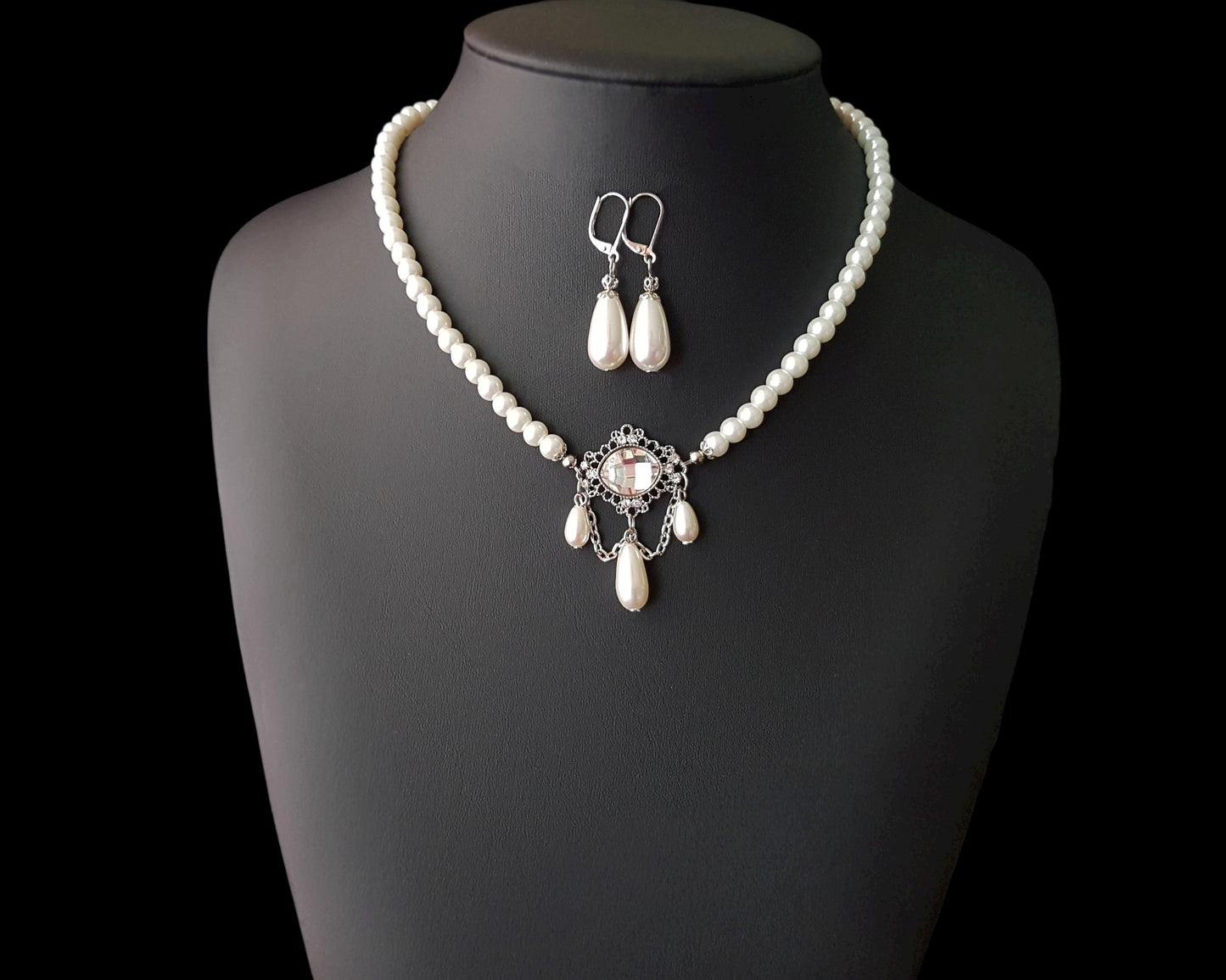Victorian Style Pearl Crystal Necklace and Earrings Set with white pearl necklace with large clear crystal and three dangling drop pearls and long back chain with another pearl drop. The matching earrings are drop shaped pearls, displayed on black. 