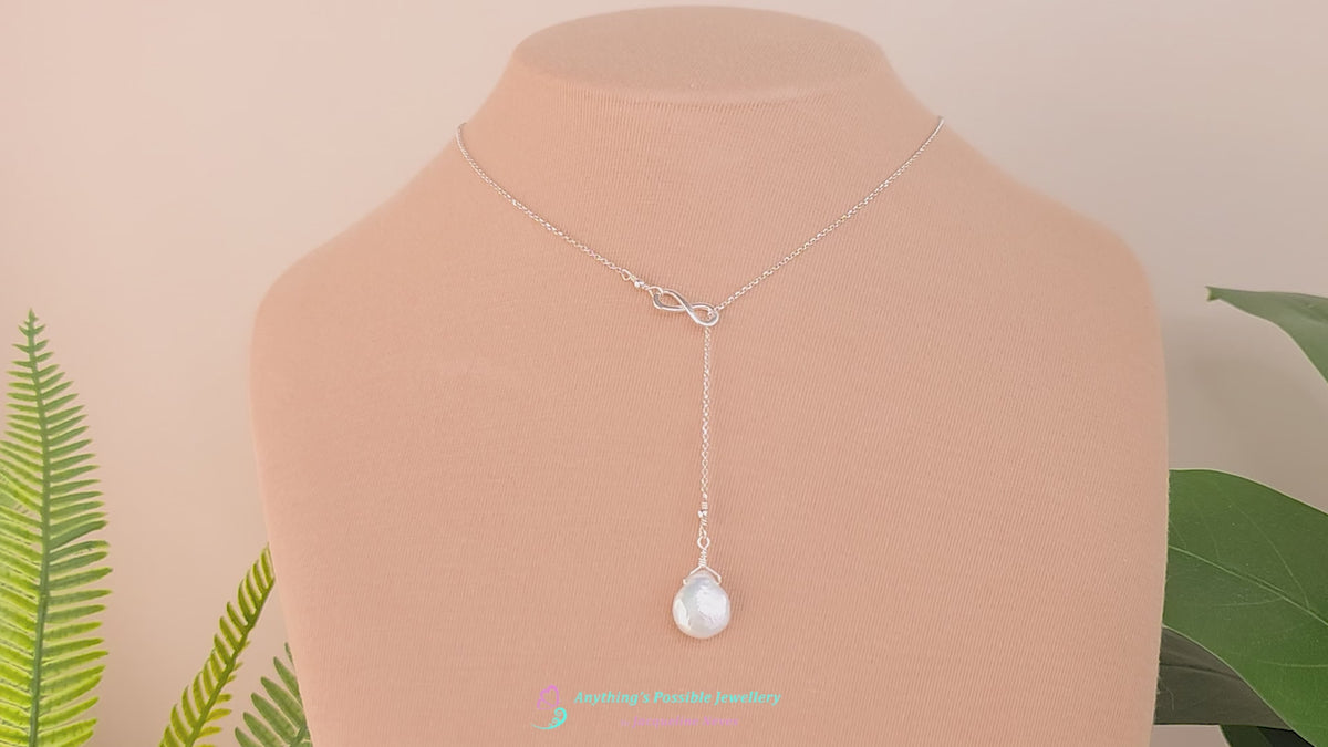 Video of a handmade large Pearl Drop Sterling Silver Infinity Lariat Necklace on beige display bust with plants in background 
