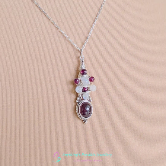 Vido of Handmade Boho style wire wrapped pendant with oval shape deep red Garnet gemstone wrapped with sterling silver wire small round garnets and faceted Moonstones on chain with beige cloth background 