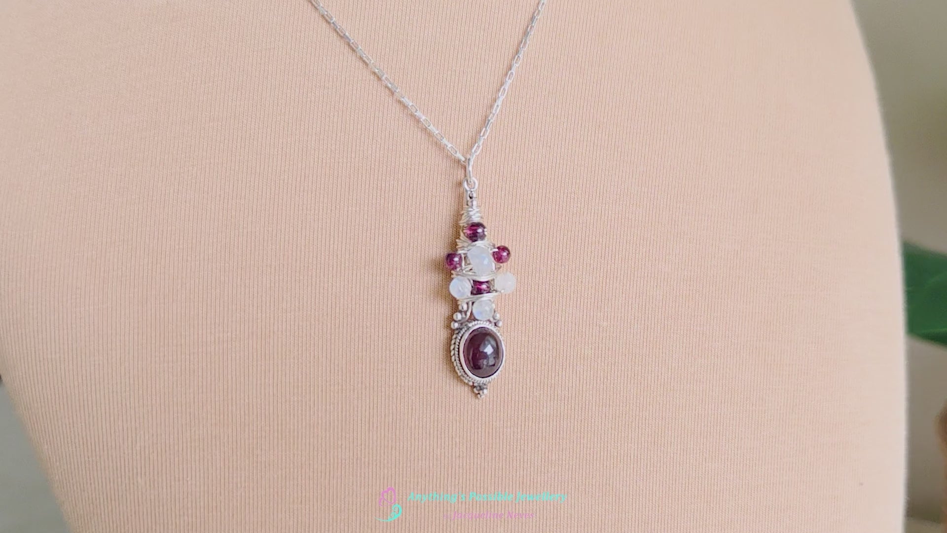 Vido of Handmade Boho style wire wrapped pendant with oval shape deep red Garnet gemstone wrapped with sterling silver wire small round garnets and faceted Moonstones on chain with beige cloth background 