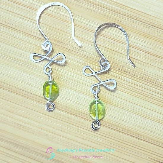 Video of handmade Silver Celtic style Peridot Dangle earrings with Eternity Coils, oval shaped green translucent Peridot stones, hand forged french hooks displayed on wood background 