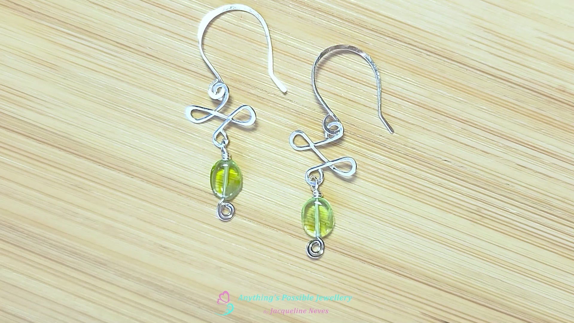 Video of handmade Silver Celtic style Peridot Dangle earrings with Eternity Coils, oval shaped green translucent Peridot stones, hand forged french hooks displayed on wood background 