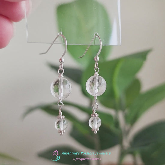 Video of A pair of clear Quartz Crystal Earrings with faceted and polished stones with Sterling Silver beads, wire wrapped and french hooks hanging with plants in the background