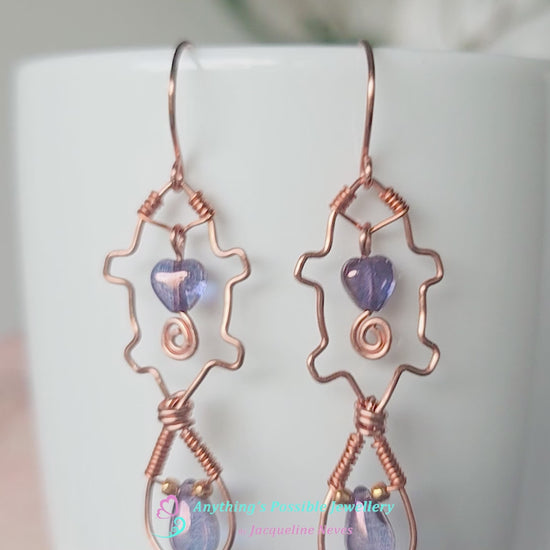 Video a pair of wire wrapped copper beaver earrings with mauve purple hearts and tear drop stones on white vase 