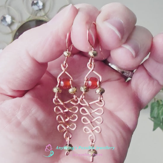Video of a pair of long handmade copper dangle earrings with a swirling Celtic river design featuring faceted red agates and sparkly faceted gold plated hematite stones on french earrings hooks on white background  