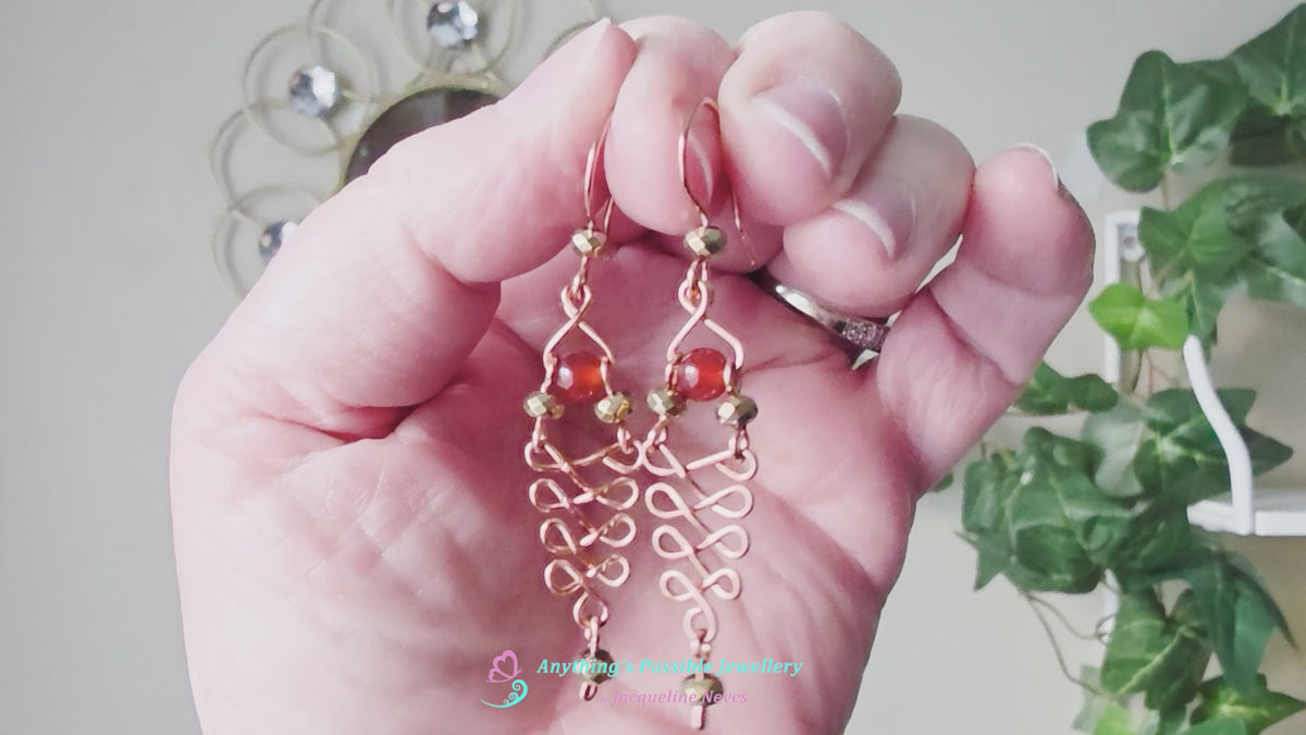 Video of a pair of long handmade copper dangle earrings with a swirling Celtic river design featuring faceted red agates and sparkly faceted gold plated hematite stones on french earrings hooks on white background  