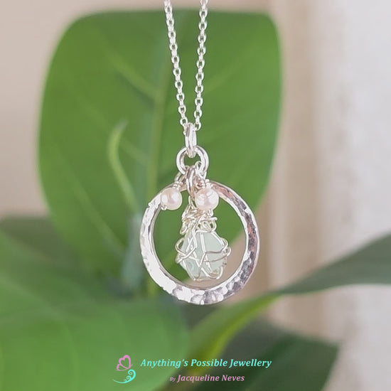 Video of Round Circle Silver Pendant Necklace with wire wrapped Aqua Blue Beach Glass and two small white pearls on chain with blurred green plant background 