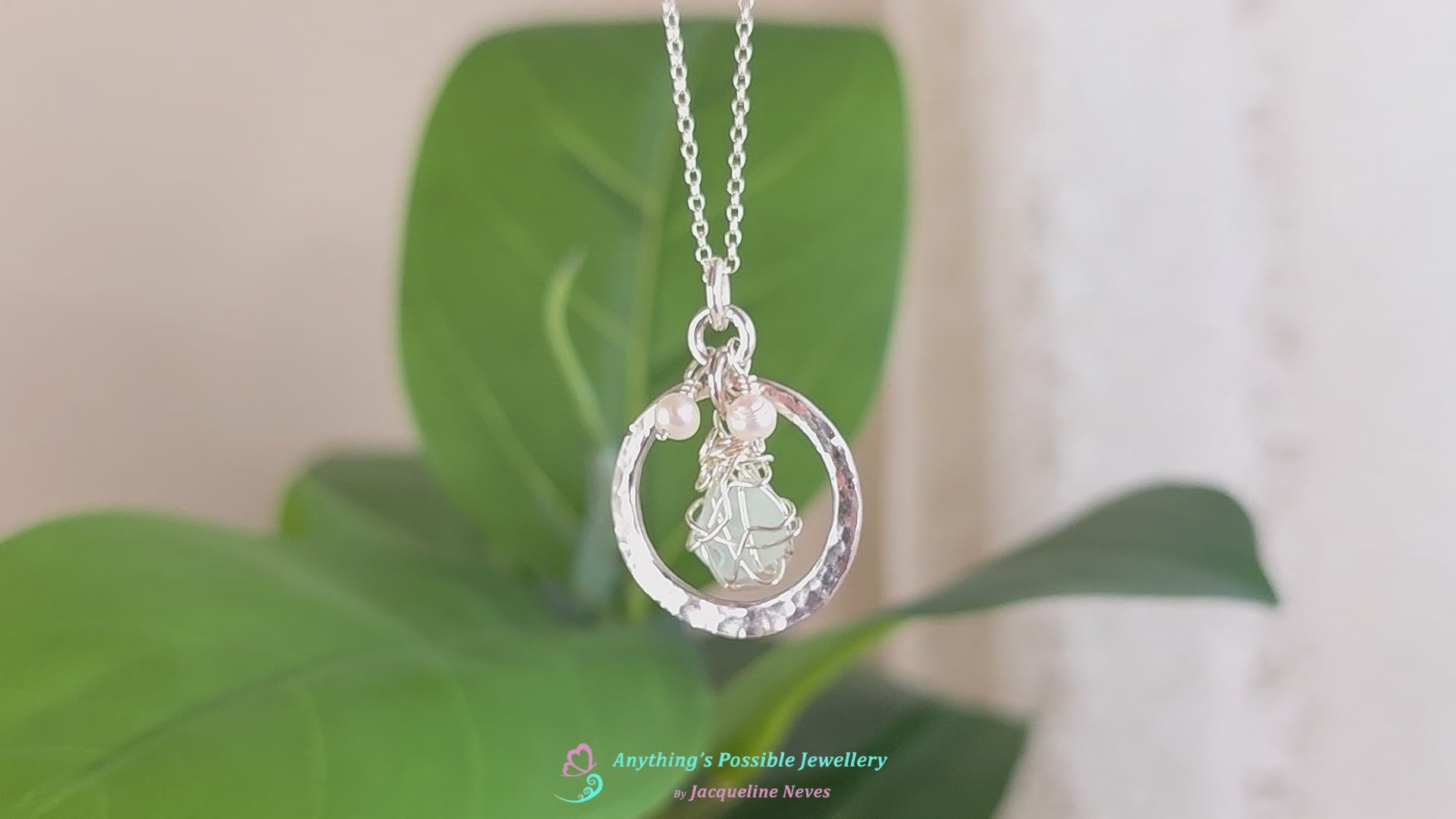 Video of Round Circle Silver Pendant Necklace with wire wrapped Aqua Blue Beach Glass and two small white pearls on chain with blurred green plant background 
