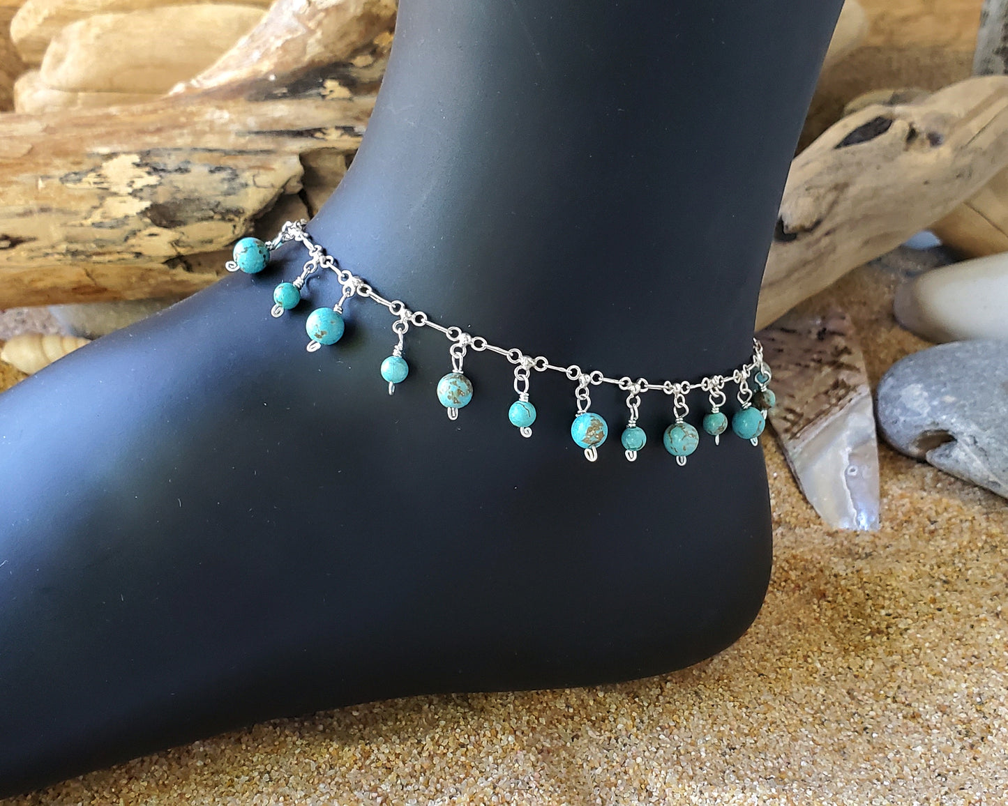 Turquoise Dangle Ankle Bracelet with many Turquoise beads dangling from a decorative silver chain. The anklet is on black foot display on sand beach with beach wood.