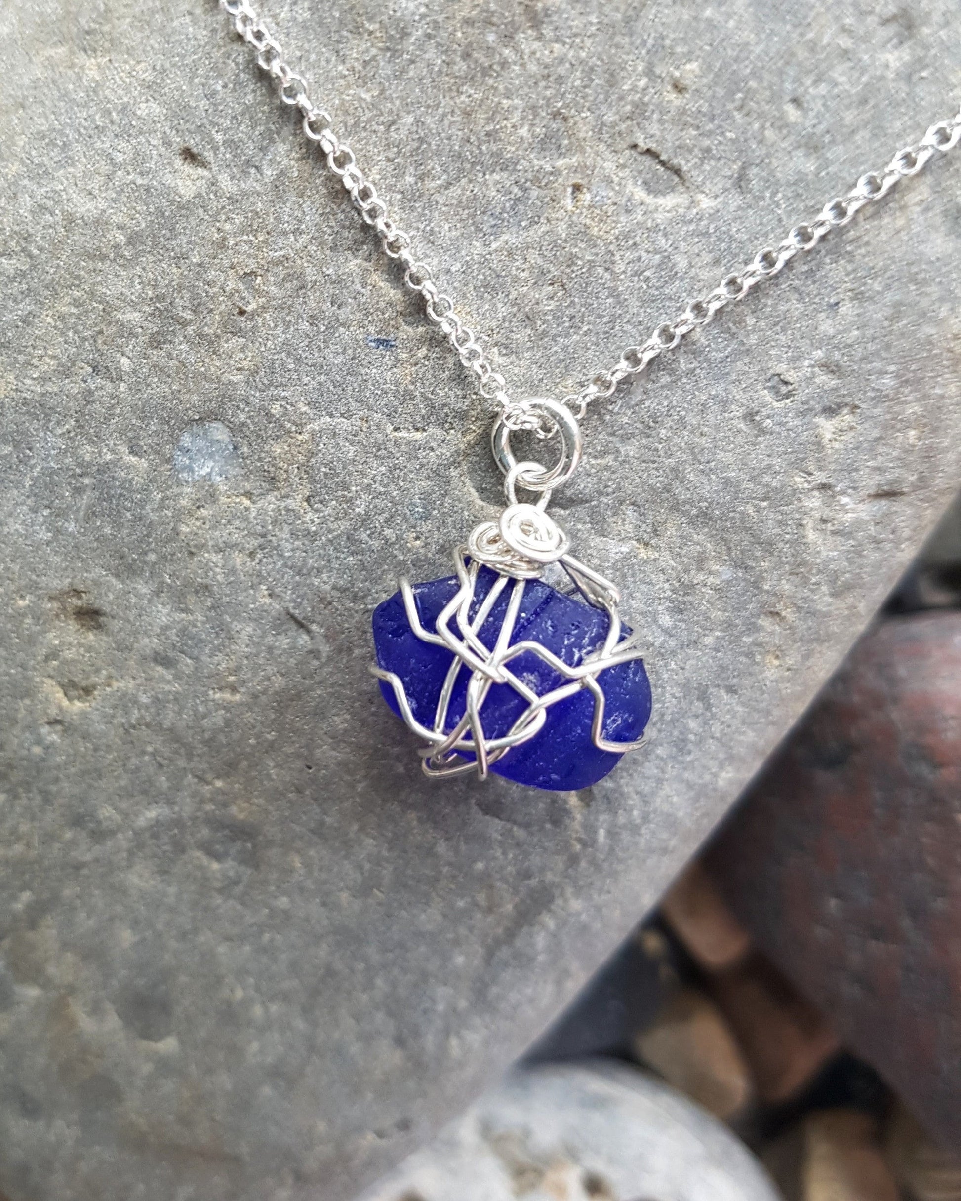 Blue Passion Sea Glass, Lake Glass-Bach GlassSapphire Blue Passion Beach Glass Pendant Necklace, Sterling Silver wire wrapped blue beach glass pendant on rolo style chain. Displayed on grey rock.