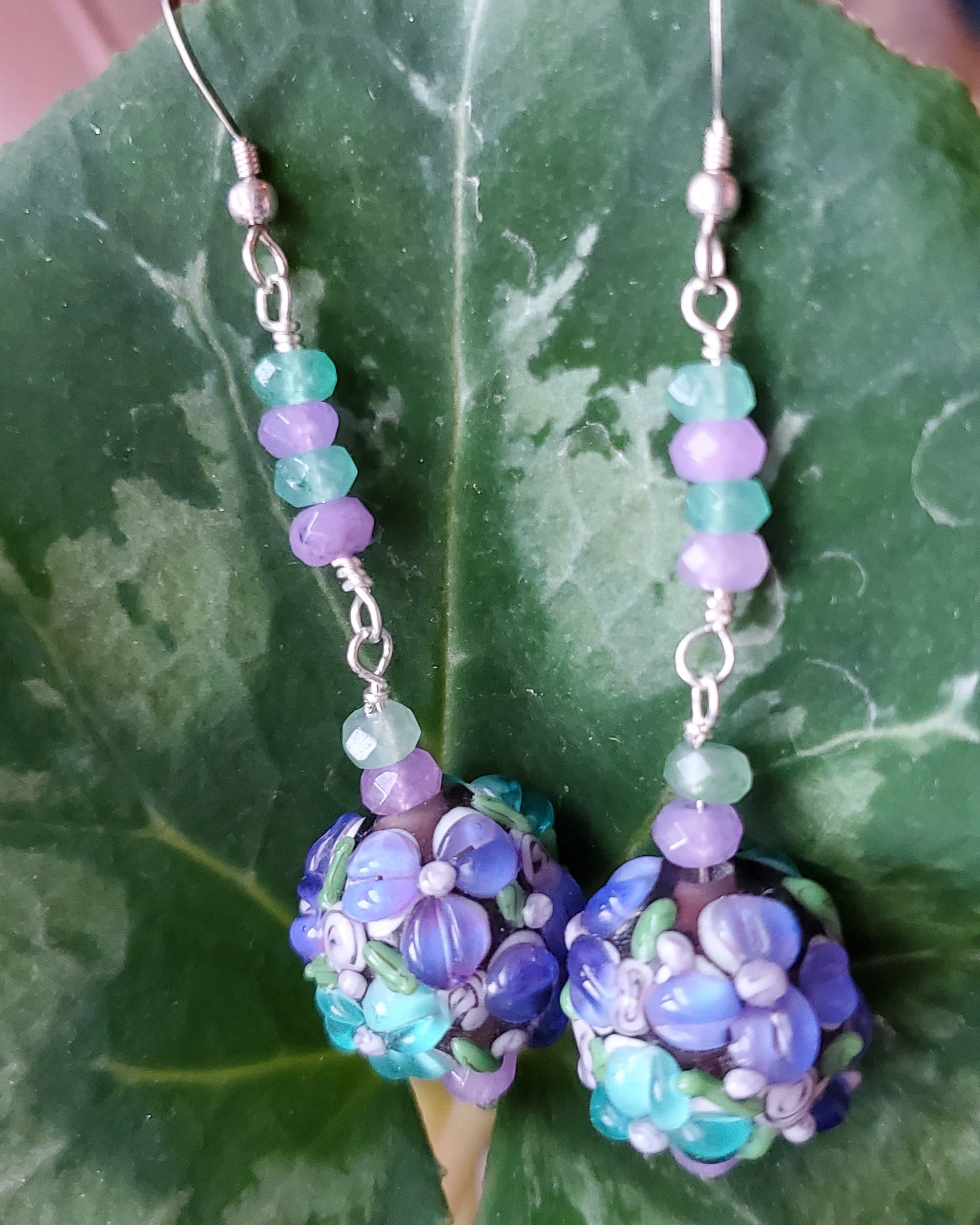 Long Lavender Flower Garden Earrings, Sterling Silver, Large round Floral Lampwork Glass beads with lavender purple and turquoise blue flowers, they dangle below a long stream of blue and lavender Quarts Stone, displayed on green Leaf