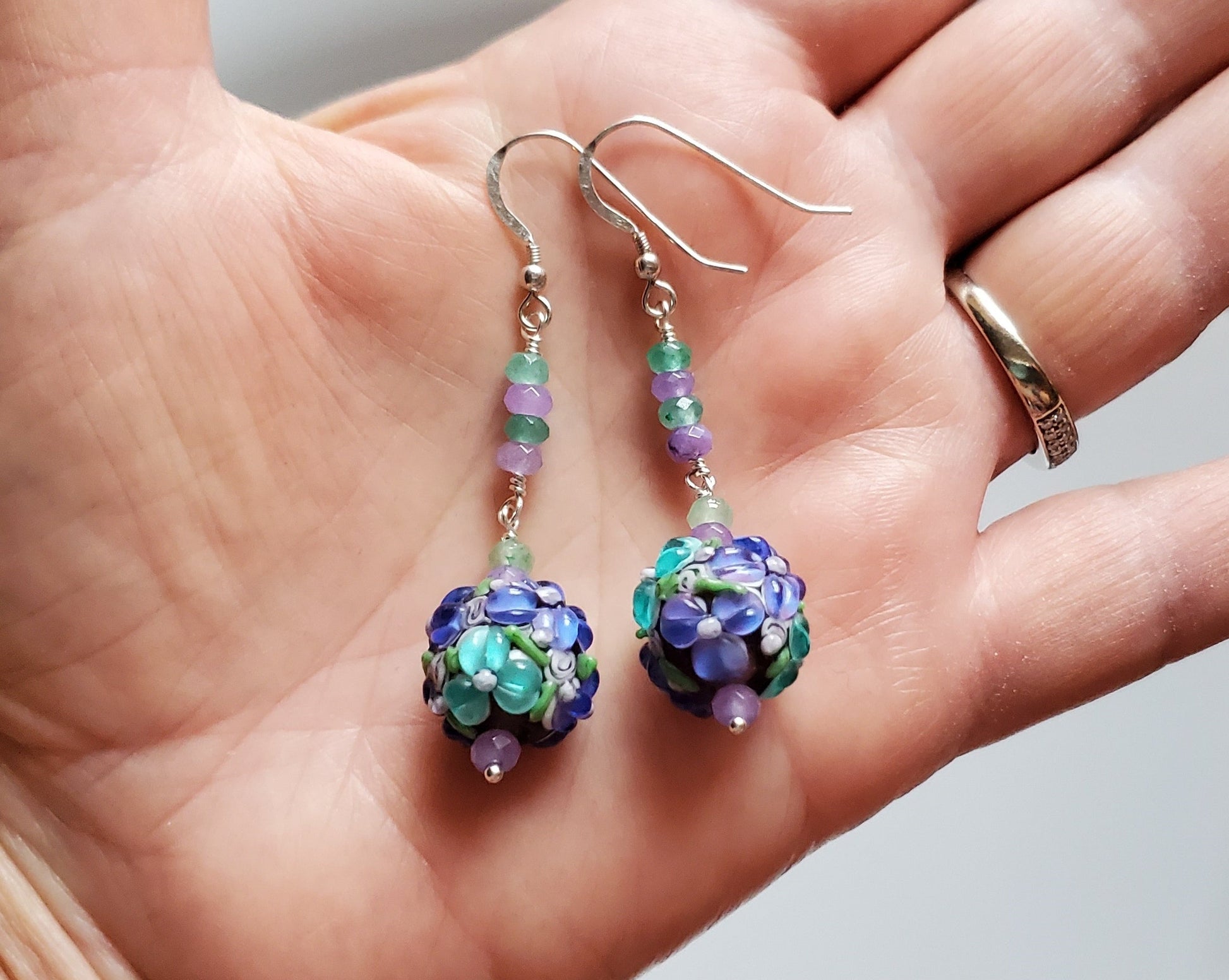 Long Lavender Flower Garden Earrings, Sterling Silver, Large round Floral Lampwork Glass beads with lavender purple and turquoise blue flowers, they dangle below a long stream of blue and lavender Quarts Stone, displayed on hand